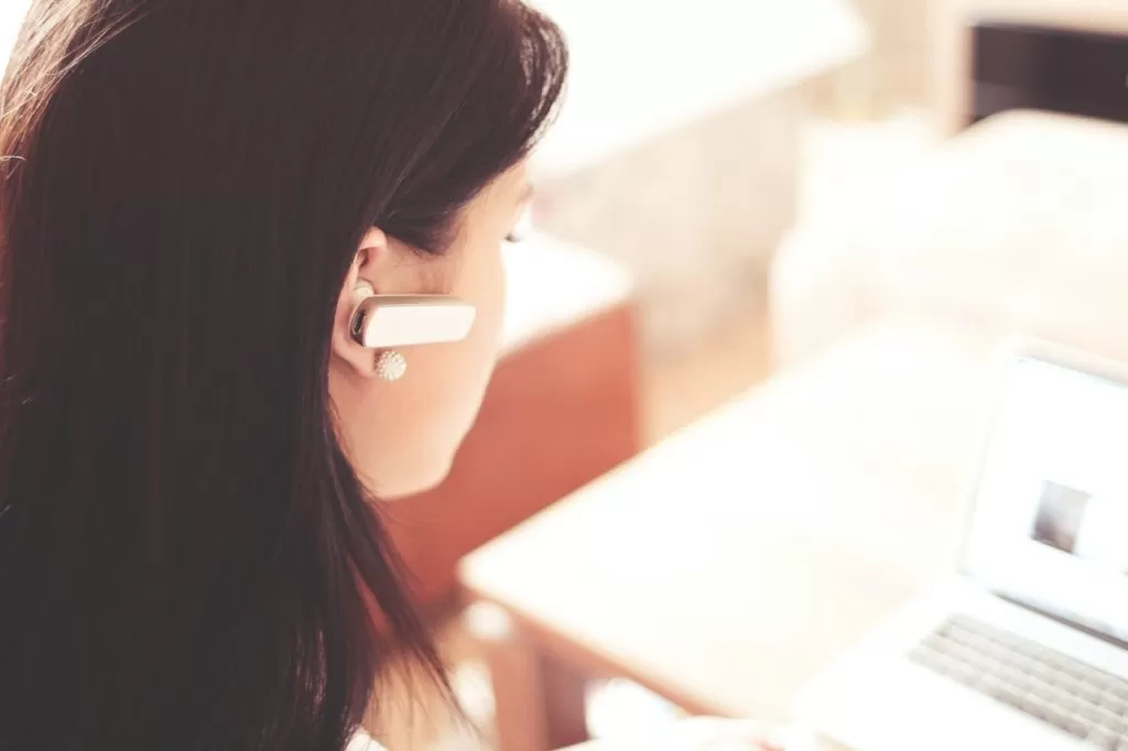 Focused woman using a headset and laptop in a sunlit room, symbolizing remote communication and digital work.