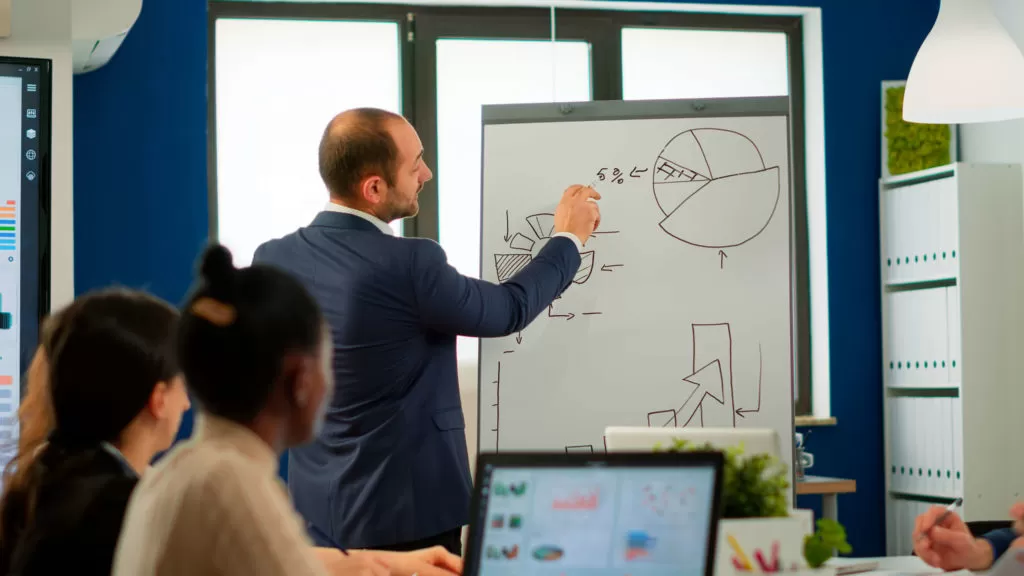 Businessman explaining pie charts on a whiteboard to colleagues in a modern office setting.